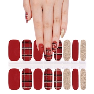 Peut inclure: Autocollants pour ongles avec différents motifs. Les autocollants comprennent des motifs unis rouges, dorés pailletés et écossais rouges avec des lignes noires et dorées. L'image montre une main avec les autocollants appliqués sur les ongles.