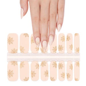 May include: A set of nail polish strips featuring a light beige base colour with a repeating gold glitter spider web pattern. The strips are arranged in rows on a backing sheet, with some applied to fingernails to demonstrate the design.