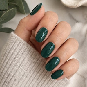 Può includere: Primo piano di una mano con smalto verde scuro a forma ovale. Le unghie sono lucide e riflettono la luce. La mano indossa un maglione beige chiaro.