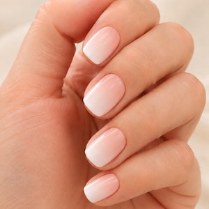 Może przedstawiać: Dłoń prezentująca manicure z gładkim efektem ombre na paznokciach. Kolor przechodzi od delikatnego, jasnoróżowego odcienia u nasady każdego paznokcia do czystej, kryjącej bieli na zaokrąglonych końcówkach, z wykończeniem o wysokim połysku.