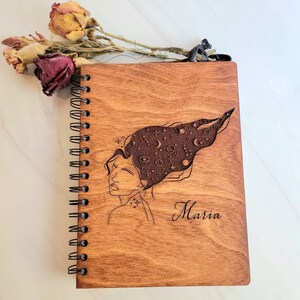 May include: A wooden journal with a spiral binding. The cover features a laser-engraved design of a woman with a galaxy in her hair. The name "Maria" is engraved below the design.