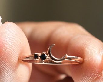 May include: A delicate gold ring with a crescent moon design and three small black gemstones. The ring is being held by a hand.
