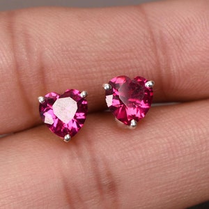 以下が含まれることがあります： ハート型のスタッドピアス。各イヤリングは、銀色の金属にセットされた鮮やかなピンク色の宝石が特徴です。宝石はファセット加工されており、光を捉えます。イヤリングは小さく繊細です。