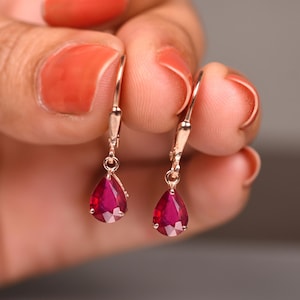 Op de afbeelding: Een paar roségouden oorhangers met peervormige, dieprode edelstenen. De oorbellen hebben een klapsluiting. De edelstenen zijn gefacetteerd en vangen het licht op.