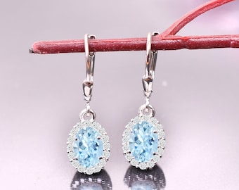 May include: A pair of silver leverback earrings. Each earring features an oval, light blue gemstone surrounded by a halo of small, clear stones. The earrings are displayed against a soft, pink background.