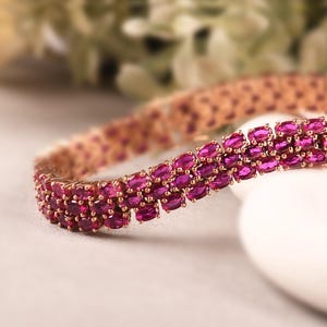 May include: A rose gold bracelet with three rows of oval-cut, deep pink gemstones. The bracelet is set on a white surface, with a blurred floral background. The jewelry piece is a statement accessory.