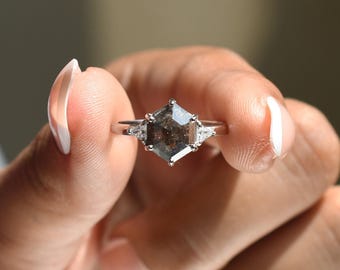 May include: A silver ring featuring a large, hexagonal, salt and pepper diamond as the centerpiece. Two smaller, triangular diamonds flank the main stone. The ring is held up, showcasing its design and sparkle.