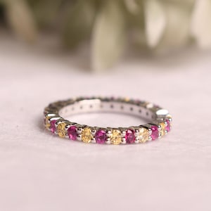 May include: A silver eternity ring featuring alternating pink and yellow gemstones. The ring is set on a light pink surface, with a blurred background of green and white foliage.