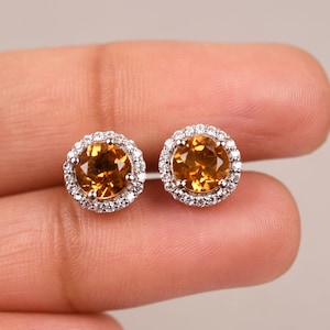 May include: A pair of gold and silver stud earrings with a round, orange gemstone surrounded by a halo of smaller, clear gemstones.