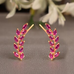May include: A pair of gold ear climbers featuring a branch-like design adorned with multiple marquise-cut vibrant pink gemstones. Each earring curves upward, mimicking the shape of a leaf-covered stem with a secure post backing.