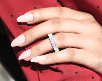 May include: A ring with a band of iridescent opal stones. The ring is on a finger with a light pink nail polish and a white tip.
