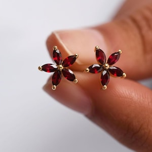 May include: A pair of gold-colored flower-shaped stud earrings. Each earring features five deep red, marquise-cut gemstones arranged around a central gold-colored bead. The earrings are held in a person's hand.