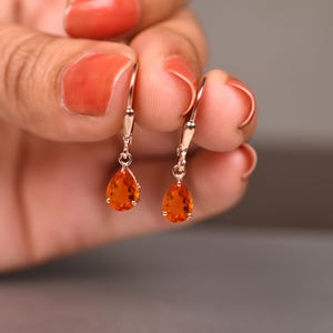 Könnte beinhalten: Ein Paar goldfarbene Klappcreolen mit tropfenförmigen, orangefarbenen Edelsteinen. Die Ohrringe werden vor einem unscharfen Hintergrund präsentiert. Die Edelsteine haben eine warme, lebendige Farbe.