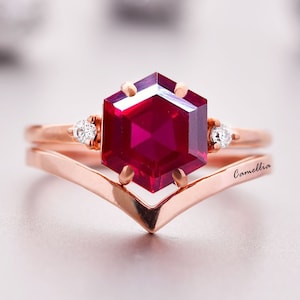 Può includere: Un anello in oro rosa con una pietra di rubino esagonale e due diamanti più piccoli. L'anello è montato su una fascia a forma di V ed è inciso con la parola "Camellia".