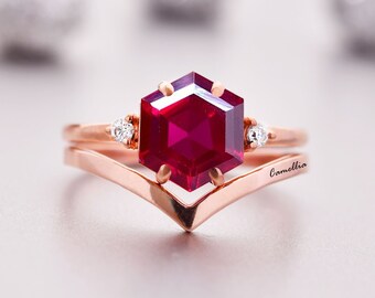 Könnte beinhalten: Ein Ring aus Roségold mit einem sechseckigen Rubin-Edelstein und zwei kleineren Diamanten. Der Ring ist auf einem V-förmigen Band montiert und mit dem Wort "Camellia" graviert.