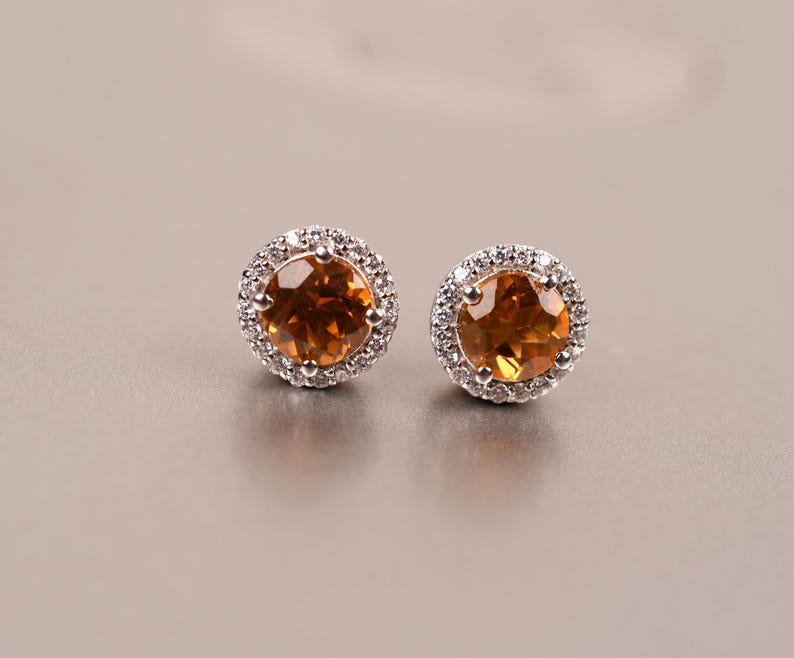 May include: A pair of silver earrings with a round, orange gemstone surrounded by a halo of smaller, clear gemstones.