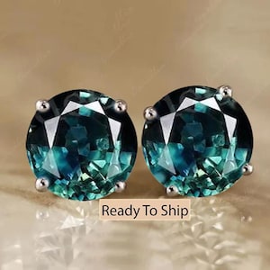 May include: A pair of round stud earrings with faceted teal and blue gemstones. The gemstones are set in silver-coloured metal. The earrings are displayed on a light brown background. The text "Ready To Ship" is visible.