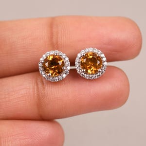 May include: A pair of gold earrings with a round, orange gemstone surrounded by a halo of smaller, white gemstones.