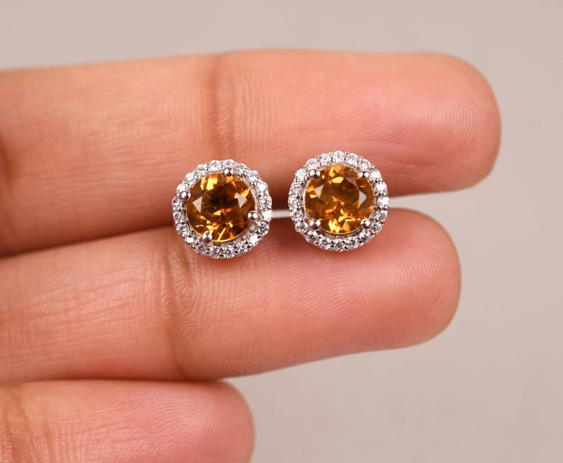 May include: A pair of gold earrings with a round, orange gemstone surrounded by a halo of smaller, white gemstones.