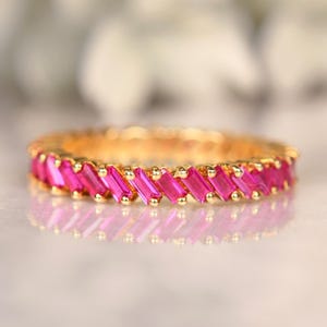 May include: A gold-toned ring with a continuous band of vibrant pink rectangular gemstones. The gemstones are set closely together, creating a solid line of color. The ring's design is simple yet elegant, suitable for everyday wear or special occasions.