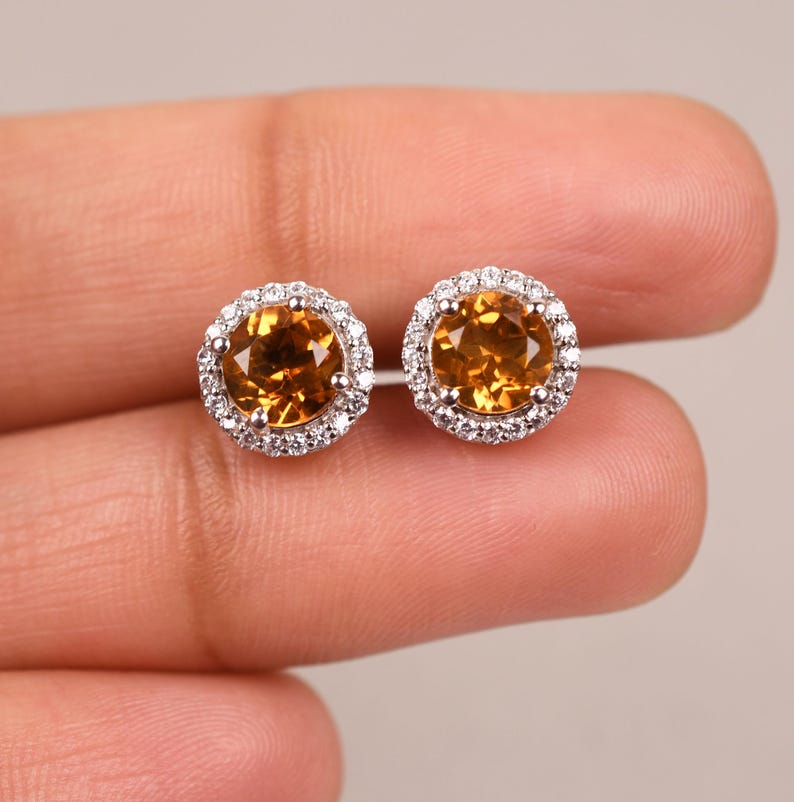 May include: A pair of gold and silver stud earrings with a round, orange gemstone surrounded by a halo of smaller, clear gemstones.