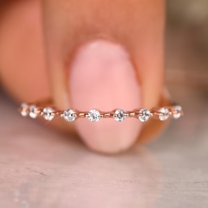 May include: A rose gold ring with a line of small, clear gemstones. The ring is held against a blurred background, highlighting the sparkle of the stones. The ring's design is simple and elegant, suitable for everyday wear or special occasions.