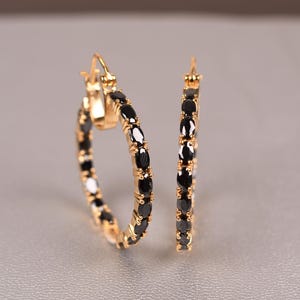 以下が含まれることがあります： 金色のフープイヤリングのペア。黒い楕円形の石がフープ全体にセットされています。