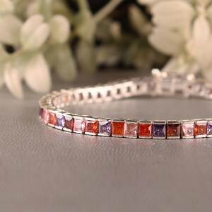 May include: A silver bracelet featuring a line of square-cut gemstones in shades of red, orange, purple, and pink. The gemstones are set closely together, forming a continuous band of colour. The bracelet is on a grey surface.