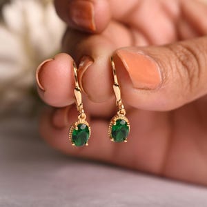 Puede incluir: Un par de pendientes dorados con cierre de palanca, cada uno con una vibrante gema verde de corte ovalado. Los pendientes se sostienen, mostrando su diseño y los intrincados detalles alrededor de las gemas.