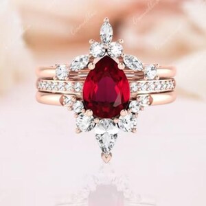 May include: A rose gold engagement ring set with a pear-shaped ruby gemstone surrounded by a halo of smaller diamonds. The ring is part of a three-piece set, with two additional bands featuring pave diamonds.