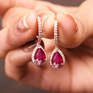 Puede incluir: Un par de pendientes de oro rosa con una piedra preciosa de rubí en forma de lágrima rodeada por un halo de diamantes más pequeños.