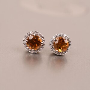 May include: A pair of silver earrings with a round, orange gemstone surrounded by a halo of smaller, clear gemstones.