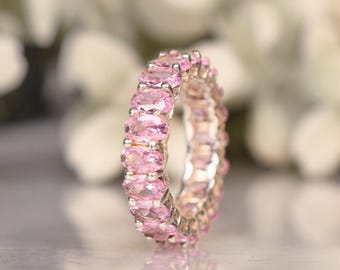 May include: A silver ring featuring a continuous band of oval-cut pink gemstones. The gemstones are set closely together, creating a full eternity design. The ring is displayed against a soft, blurred background.