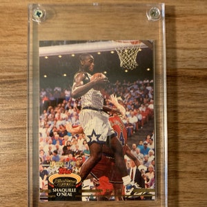 May include: A 1992-93 Topps Stadium Club basketball card featuring Shaquille O'Neal in a white and blue Orlando Magic uniform, with a gold border and a gold "Stadium Club" logo.