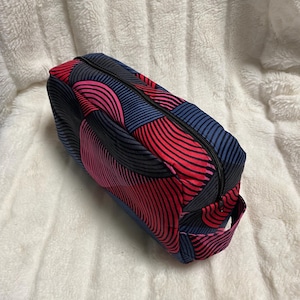 Op de afbeelding: Rechthoekige make-uptas met een levendig patroon van rode, roze en marineblauwe gebogen lijnen. De tas heeft een zwarte ritssluiting en een klein handvat. Geplaatst op een witte, zachte ondergrond.
