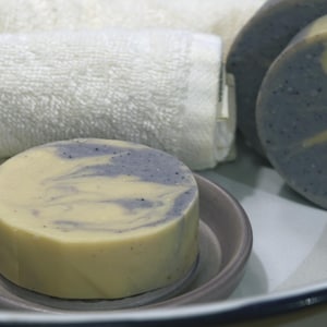 May include: A close-up of a round, swirl-patterned bar of soap with a light yellow and purple color scheme. The soap is sitting on a gray dish.