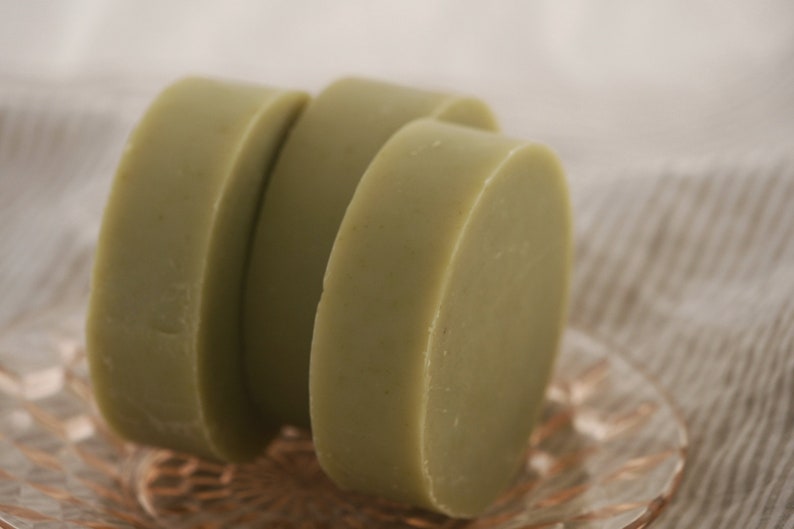 K&ouml;nnte beinhalten: Drei runde St&uuml;cke gr&uuml;ner Seife, die &uuml;bereinander auf einem rosa und durchsichtigen Glasteller gestapelt sind.