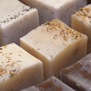 以下が含まれることがあります： 正方形の手作り石鹸のクローズアップ。石鹸は明るい色で、上部にハーブや種子が見えるものもあります。石鹸は互いに近くに配置されており、自然な質感と成分を示しています。