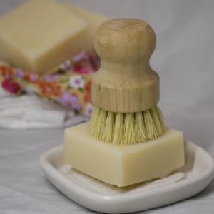 May include: A light-coloured soap bar sits on a white, leaf-shaped dish, with a wooden-handled brush on top. The brush has light-coloured bristles. Additional soap bars and a floral patterned cloth are in the background. The items are likely for cleaning.
