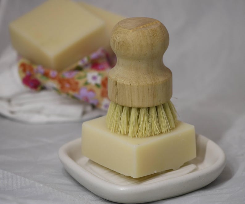 May include: A light-coloured soap bar sits on a white, leaf-shaped dish, with a wooden-handled brush on top. The brush has light-coloured bristles. Additional soap bars and a floral patterned cloth are in the background. The items are likely for cleaning.