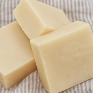 May include: Three square bars of pale yellow handmade soap, arranged on a striped fabric. The soaps have a smooth surface and a clean appearance. The image suggests a natural skincare product, possibly for the body.