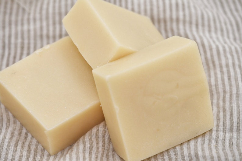 May include: Three square bars of pale yellow handmade soap, arranged on a striped fabric. The soaps have a smooth surface and a clean appearance. The image suggests a natural skincare product, possibly for the body.