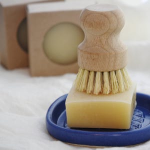 May include: A wooden handled dish brush with natural bristles rests on a square bar of soap in a small blue ceramic dish. Two round soaps are in cardboard boxes in the background. The items are on a white cloth.