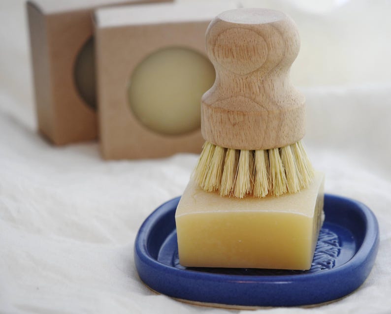 May include: A wooden handled dish brush with natural bristles sits on a square bar of soap in a small blue ceramic dish. Two round soaps are in cardboard boxes in the background. The items are on a white cloth.
