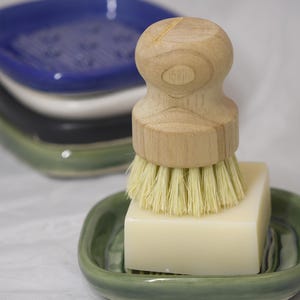 May include: A wooden handled dish brush with light bristles rests on a bar of soap in a green ceramic soap dish. Stacked behind are three additional ceramic soap dishes in blue, white, and black.