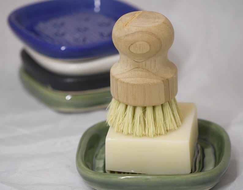 May include: A wooden handled dish brush with light bristles rests on a bar of soap in a green ceramic soap dish. Stacked behind are three additional ceramic soap dishes in blue, white, and black.