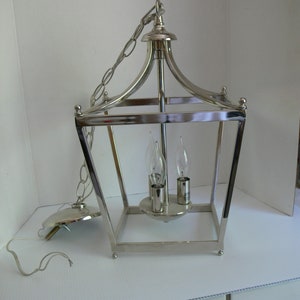May include: A silver-toned, rectangular lantern-style light fixture with a chain for hanging. The fixture has a decorative top and holds three candelabra-style bulbs. The fixture is designed for indoor use.