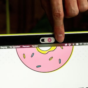Könnte beinhalten: Ein Laptop mit einem rosafarbenen und gelben Donut auf dem Bildschirm. Der Laptop hat eine Webcam-Abdeckung über der Kamera. Der Bildschirm zeigt auch die Wörter "Geçmiş", "Yer İşaretleri", "Pencere" und "Yardım".