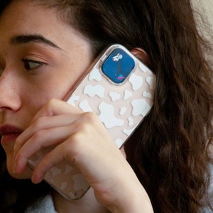 以下が含まれることがあります： 牛柄の白とピンクの携帯電話ケース。ケースは透明な背面とカメラレンズに小さな宇宙飛行士のステッカーが付いています。