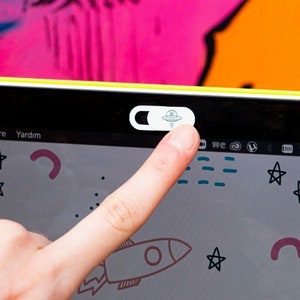 May include: A white laptop webcam cover with a UFO design. The cover is being applied to a laptop screen with a finger.